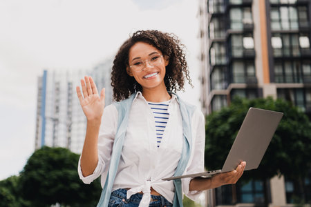 Smiling woman with laptop waves at camera in sunny urban city scene showcasing casual fashion and modern work vibe outside stylish buildingsの写真素材