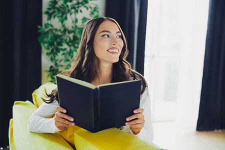 Happy woman reads a book on a bright yellow couch at home in a cozy living room creating warm relaxed vibesの写真素材