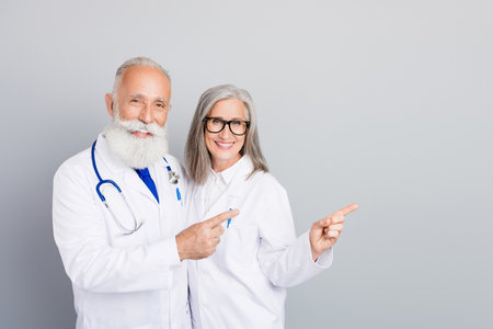 Two experienced doctors and a colleague pair in white coats smile and point to a message in a friendly medical scene of care and consultationの写真素材