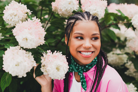 Portrait of a young woman with braids in a pink jacket among hydrangeas smiling in a sunny garden setting for lifestyle and fashionの写真素材