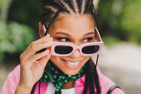 Young woman with pink sunglasses smiles outdoors wearing braids and a green scarf in a sunny park settingの写真素材