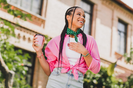 Happy young woman with braids wearing a pink jacket and green scarf holds a coffee cup smiles on a sunny street scene in a vibrant urban neighborhoodの写真素材