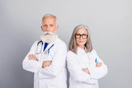Two experienced doctors pose with arms crossed in white coats against a grey background showcasing medical consultation and professional healthcare teamの写真素材