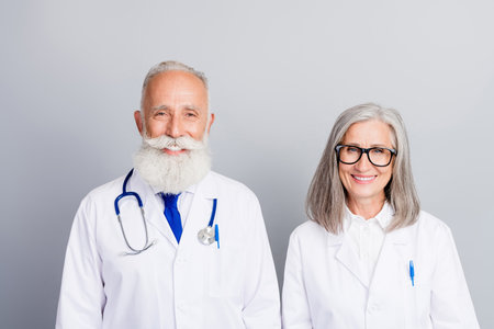 Senior male doctor and senior female physician smiling in white coats with stethoscope ready for consultation in a bright clinic settingの写真素材