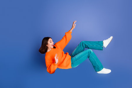 Young woman in casual orange sweater and teal jeans appearing to float, expressing excitement on a blue backgroundの写真素材