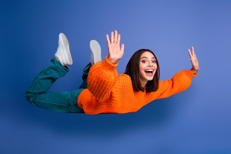 Cheerful young woman in orange sweater smiling brightly and posing freely against blue background showcasing casual funの写真素材