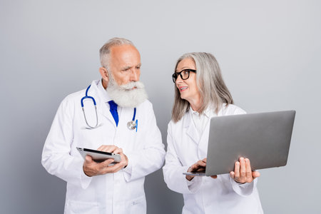 Two experienced doctors discuss patient care using tablet and laptop during a professional consultation in a clinical settingの写真素材