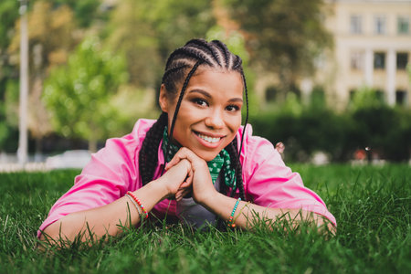 Young woman with braids smiles lying on grass wearing a pink jacket in a sunny park with an urban city backdropの写真素材