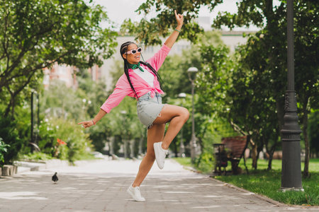 A stylish young woman in a pink shirt denim shorts and white sneakers leaps in a sunny urban park for a playful street style momentの写真素材