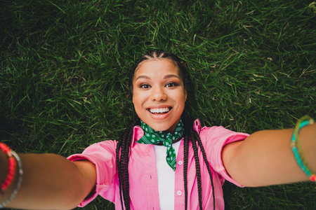 Young woman with braids lying on grass smiling for a cheerful outdoor selfie wearing a pink shirt and green bandanaの写真素材