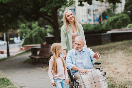 Lulfamily stroll in park with grandpa in wheelchair and girl smiling while mother watches and friends share care love and joy in a sunny day in the parkの写真素材