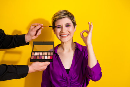 Young woman smiles as makeup is applied by another person against a vibrant yellow backdrop showcasing glamour chic style fashion and beautyの写真素材