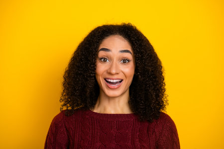 Cheerful young mixed race woman with curly hair wearing burgundy sweater smiles against bright yellow background for lifestyle fashionの写真素材