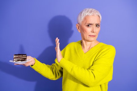 Elderly woman in bright yellow sweater refuses chocolate cake with surprised look in studioの写真素材