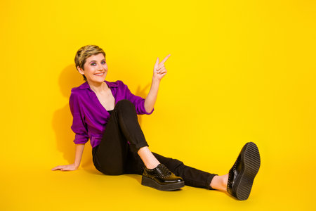 Young female model sits on yellow background wearing a purple blouse and black pants smiling and...