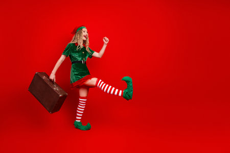 Festive young woman dressed as a Christmas elf joyfully posing in an exuberant outfit on a vibrant red backdropの写真素材