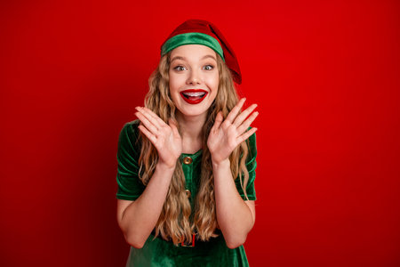 Excited young woman dressed as a festive elf posing against a red background, showcasing holiday cheer and enthusiasm.の写真素材