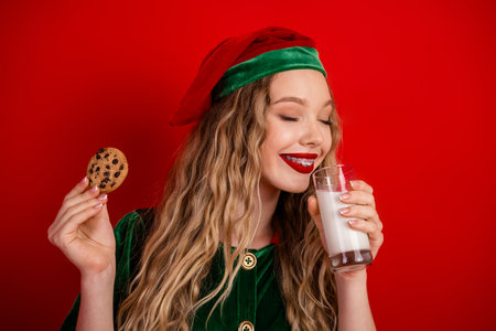 Merry Christmas elf enjoying a delicious treat of milk and cookies against a vibrant red holiday-themed backgroundの写真素材