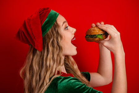Young woman dressed as a festive elf enjoys a delicious hamburger against a vibrant red background with holiday vibesの写真素材