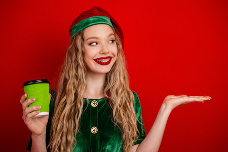 Smiling festive girl in elf costume holding coffee and gesturing, posing against red background for...