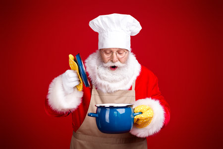Santa the chef in a red holiday kitchen prepares a Christmas feast with a blue pot and plates while smiling and cookingの写真素材