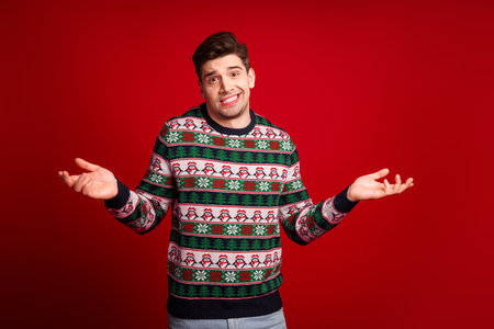 Funny cheerful man in a Christmas sweater shrugs playfully against a bold red background for holiday marketing and festive promotion ideasの写真素材