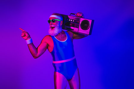 Energetic senior man in retro blue jumpsuit dances with a boombox under purple neon lights for fitness party lifestyleの写真素材
