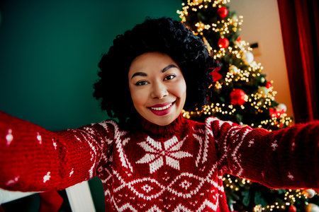 Joyful woman in red festive sweater taking a selfie by the beautifully decorated Christmas tree during the holidaysの写真素材