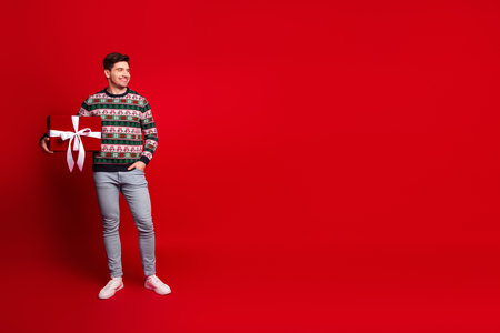 Cheerful man with gift posing in a bright red studio celebrates Christmas with a festive sweater and big smile for holiday shoppers and warm seasonal vibesの写真素材