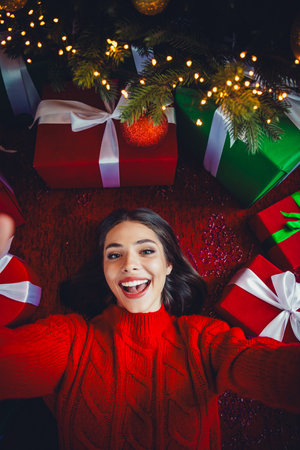 Joyful woman in a bright red sweater lies on the floor under a festive tree surrounded by gifts and...
