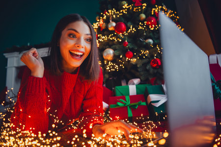 Playful holiday moment as a happy woman in a red sweater smiles at a glowing Christmas tree and gifts during a cozy home setupの写真素材