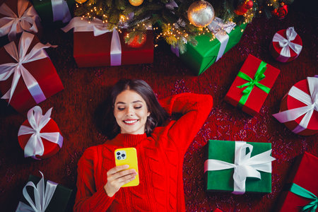 Cheerful festive moment as a woman in a red sweater relaxes on the floor among Christmas gifts and a...