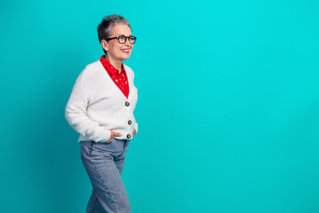 Smiling elderly woman in white cardigan red blouse and blue jeans against teal background showcasing casual fashion style for holiday seasonの写真素材