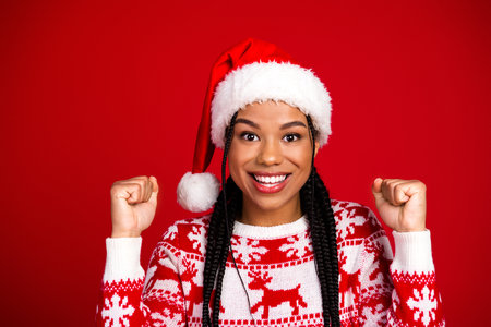 Cheerful multiethnic woman in a Santa hat and red Christmas sweater celebrates the holidays with fists up against a bold red backgroundの写真素材
