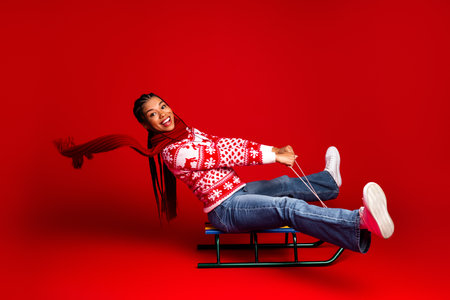 Funny winter joy as a young girl on a sled laughs across a red studio background wearing a festive...