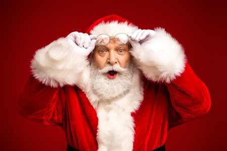 Santa Claus in a red suit with white fur surprises with wide eyes while adjusting glasses in a festive Christmas portrait for holiday campaignsの写真素材
