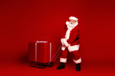 Santa pulls a large red gift on a sleigh beside a bold red background for a festive Christmas scene full of joy and holiday magicの写真素材