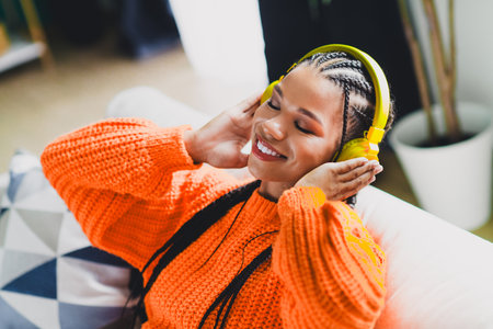 Woman in bright orange knit sweater relaxing at home listening to music with yellow headphones on a...