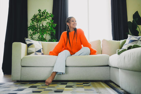Bright living room scene with a young woman in an orange sweater relaxing on a sofa at home...