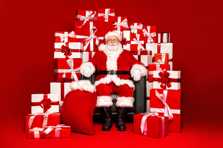 Santa Claus sits in a red room surrounded by wrapped gifts and a large sack of presents for a festive family moment and holiday shopping dreamの写真素材