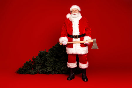 Santa in a red suit with white fur stands with an axe beside a Christmas tree against a red background creating a festive season moodの写真素材