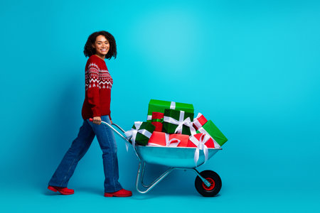 Playful woman pushes a blue wheelbarrow full of colorful gifts and wrapping presents celebrating Christmas season with joy and holiday magic in a bright studioの写真素材