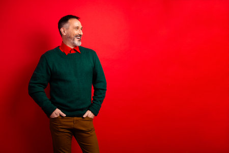 Cheerful mature man in green sweater smiles against red background ready for holiday shopping and festive funの写真素材