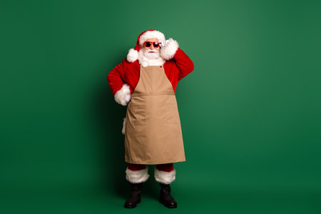 Santa in red costume with beige apron stands on a green background wearing hat and sunglasses ready for festive funの写真素材