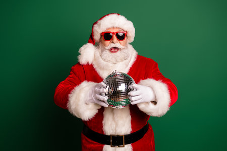 Santa in red suit holds a disco ball in a festive portrait for Christmas promotions on a green backgroundの写真素材
