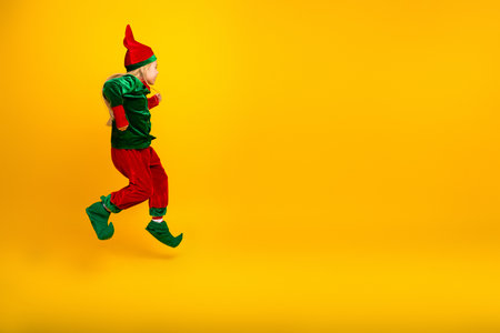 Cheerful young girl dressed as an elf jumping in excitement against a vibrant yellow background, spreading holiday joyの写真素材