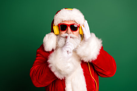 Santa in red suit with yellow headphones on green background posing with finger to lips for stock photoの写真素材