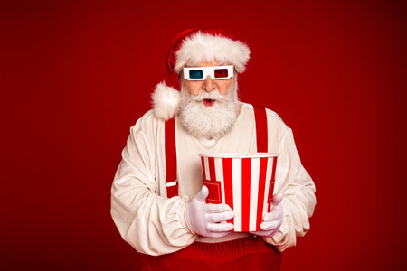 Santa in red suit holds striped popcorn bucket wearing 3d glasses against a bold red background for a festive christmas seasonの写真素材