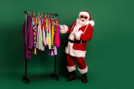 Santa in a festive red suit poses beside a colorful clothing rack full of bright outfits against a green background perfect for holiday fashion and seasonal shopping promotionsの写真素材