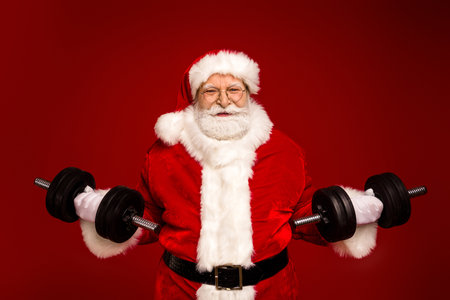 Santa lifts heavy dumbbells in a festive red studio for a humorous fitness themed Christmas image for holiday promotionsの写真素材